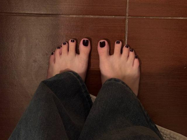 Os pés descalços de MonaTrio, com as unhas dos pés pintadas de preto, estão em pé sobre um chão de azulejos marrons, vestindo calças jeans azul-escuro, mostrando uma leve sombra sob os dedos dos pés..