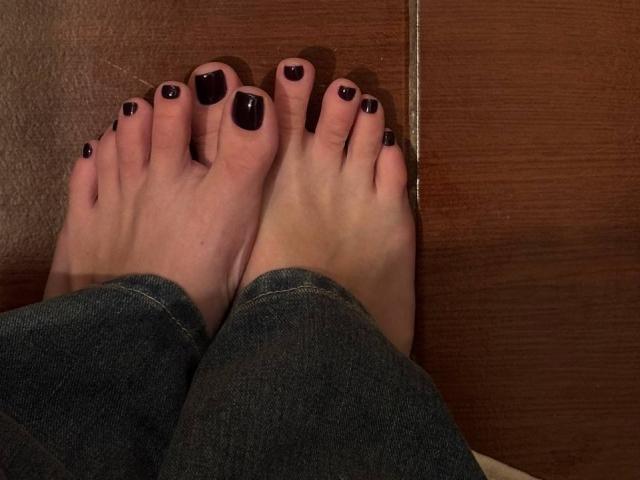 MonaTrio's feet with black-painted toenails, wearing blue jeans, resting on wooden floor. Her toes are slightly curled, showing clean, well-maintained feet.