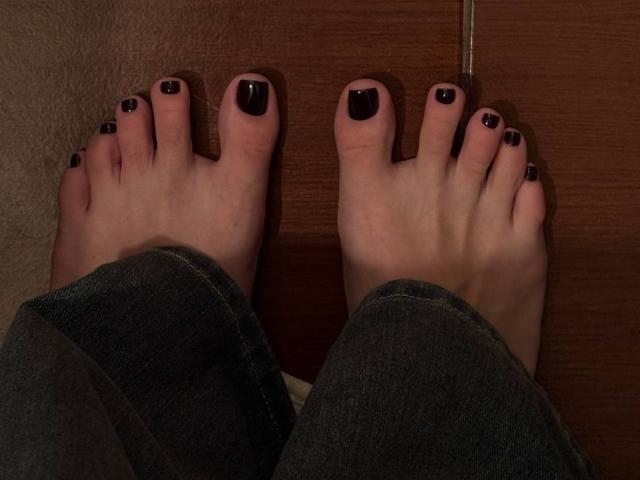 Los pies de MonaTrio, con las uñas de los dedos pintadas de negro, están sobre un suelo de madera, y lleva unos vaqueros de mezclilla oscuros. Sus dedos están ligeramente separados, mostrando claramente las uñas pintadas..
