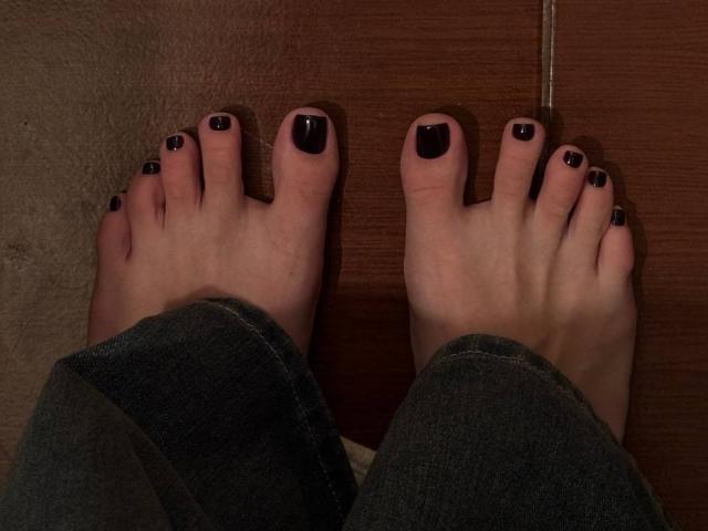 De voeten van MonaTrio, met donkerpaarse nagellak, met gespreide tenen, draagt donkergroene jeans en staat op een houten vloer. De zichtbare tenen zijn groot en lichtelijk vuil..