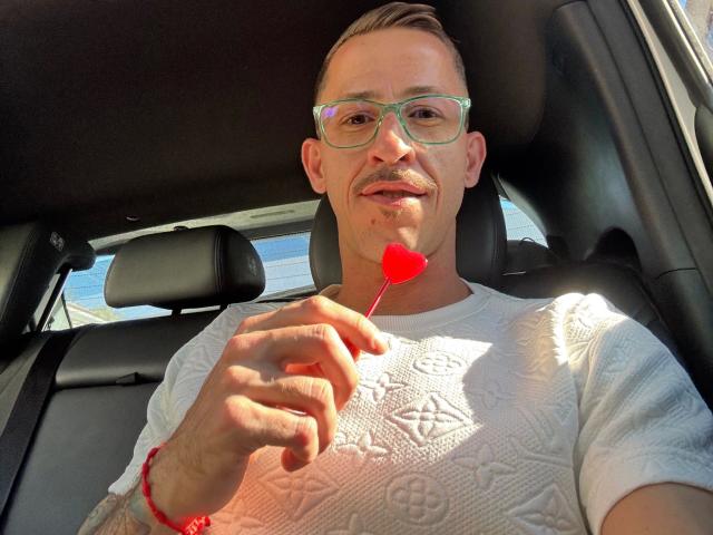 LucasJandre-hot with glasses, white textured shirt, red bracelet, holding red heart lollipop, smiling, mustache, sunlit car, short brown hair, light skin, casual selfie.