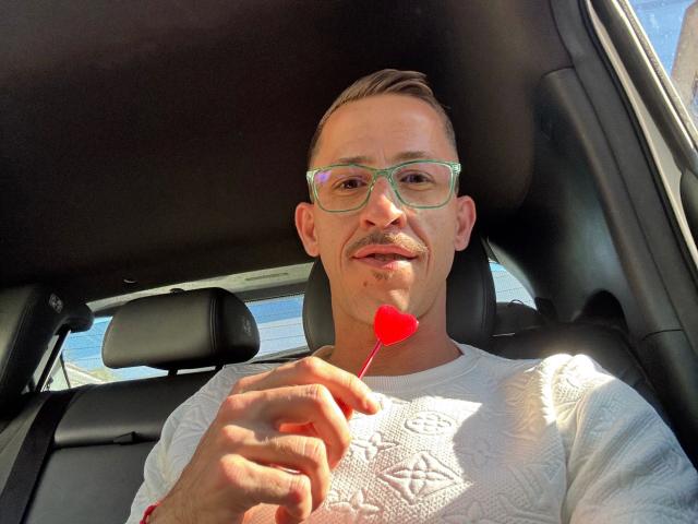 LucasJandre-hot, a white man with short brown hair and a mustache, wears green glasses and a white textured shirt. He holds a small red heart-shaped lollipop near his mouth. Sunlight illuminates his face and chest. The car's black interior is visible, with a seat and headrest in the background. The image is bright and detailed, capturing LucasJandre-hot's relaxed expression and the lollipop's vivid red color.