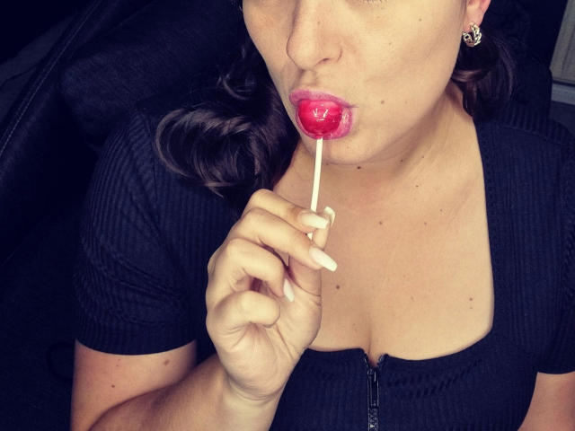 SiegruneValkyra-hot, with fair skin and brown wavy hair, holds a red lollipop to her slightly parted lips. She wears a black ribbed shirt with a zipper, showing cleavage. Her right hand, with long, pale nails, grips the white stick. She has small earrings. The image is a close-up, focusing on her face and upper chest. The lollipop's glossy texture contrasts with her matte shirt. The photo has a slight blue tint.