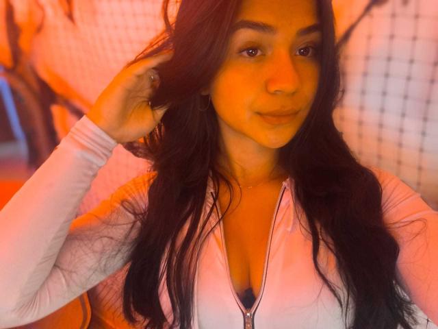 RouxiMori with long black hair, wearing a white zip-up top, showing her small tits, has a subtle smile, orange light on her face, and hand in her hair.