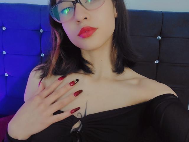 CamiVargas with dark hair, red lipstick, glasses, off-shoulder black top, red nails, hand on chest, pale skin, blue headboard, and black tufted background.