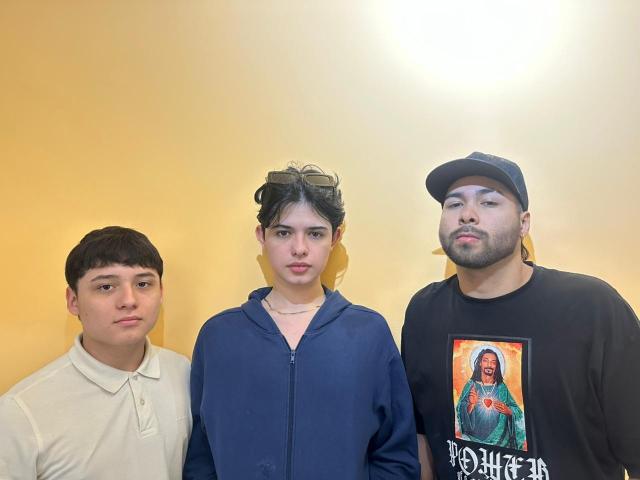 Three people stand in a row, centered in the image. HotThreesoome on the left has short brown hair and wears a white polo shirt. HotThreesoome in the middle has dark hair in a ponytail with sunglasses on top, wearing a blue hoodie. HotThreesoome on the right wears a black cap and a black t-shirt with a colorful graphic of a woman in a green traditional outfit. The right HotThreesoome has a beard and a serious expression. The background is a plain yellow wall.