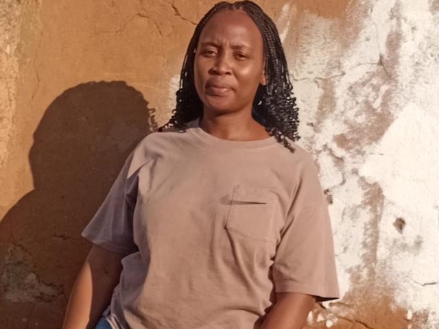 BigbootyCurvesMilf with dark brown skin, medium build, wearing a beige t-shirt, and braided hair, standing against a rough wall, looking slightly annoyed. Her tits are small.