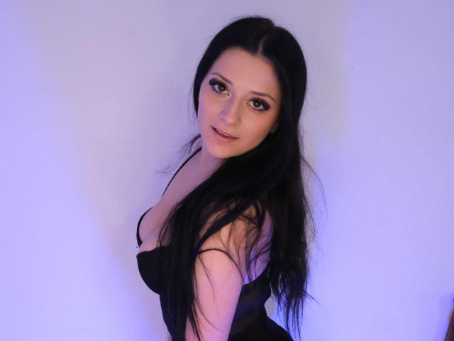 MissMayy with long black hair, looking up, wearing a black dress, showing her small tits, pale skin, and brown eyes, against a purple-lit background.