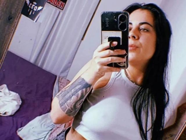 SindyLove with long black hair, wearing a white crop top, showing off her tits and tattooed arm, taking a mirror selfie with a smirking face.