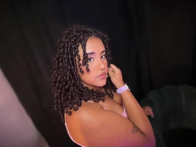 MelodyDubois with curly black hair, pink bra, and purple wristband, looking over her shoulder with a slight smile, touching her cheek. Her tits are visible, showing a small tattoo.
