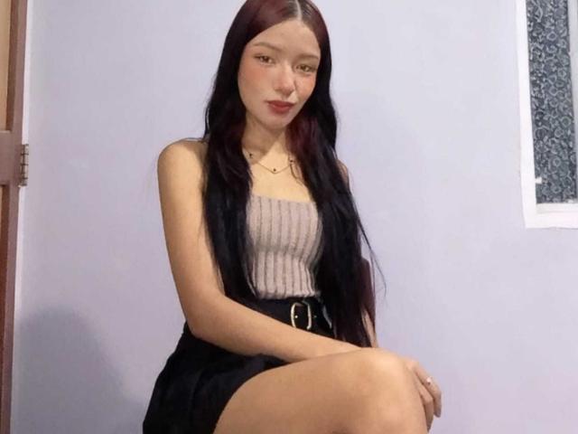 IvorySelene has long dark red hair, light skin, and wears a strapless gray top with a black skirt and belt. She's sitting with one leg crossed.