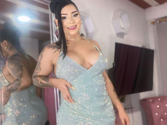 DioSaily con grandi seni, vestita con un abito azzurro chiaro scintillante, braccio tatuato, capelli neri raccolti in una coda di cavallo, sorridente, in piedi di fronte a uno specchio, stanza rosa..