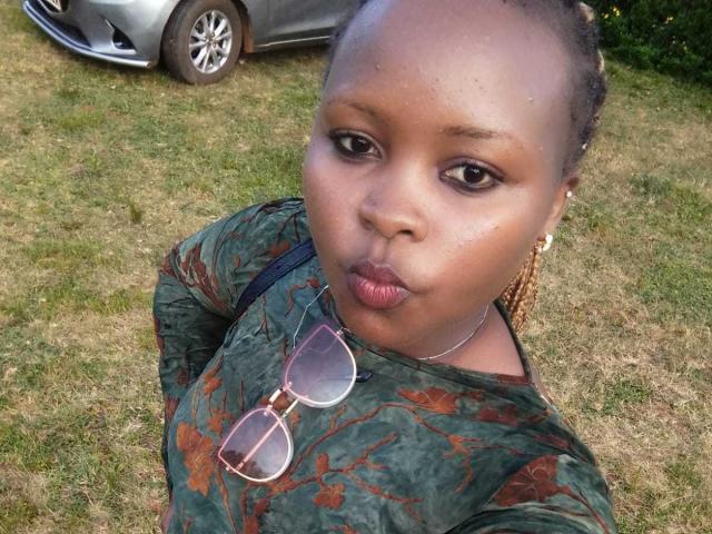 Pinacol, a Black woman with a medium brown skin tone, stands outdoors on a grassy lawn. She has braided hair pulled back and wears a green, floral-patterned long-sleeve top. She has dark brown eyes and full lips with a slight pout. She wears round, pink-tinted eyeglasses on a black strap and has a thin, silver necklace. The silver car in the background has a visible front and partial side. The grass is green with some yellow patches. The image has a casual, selfie-style composition.