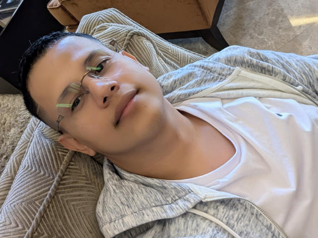 Close-up of LiamB, a young man with light brown skin and short black hair, wearing clear glasses with green-tinted lenses. He is lying on a beige, textured chair, wearing a gray, zip-up hoodie over a white t-shirt. LiamB's face is slightly tilted upward with a relaxed, subtle smile. His eyebrows are thin and dark, and he has a small, slightly pointed nose. The background includes a wooden chair and a patterned floor. The light is natural, highlighting his skin's smooth texture. The composition focuses on LiamB's upper body and face.