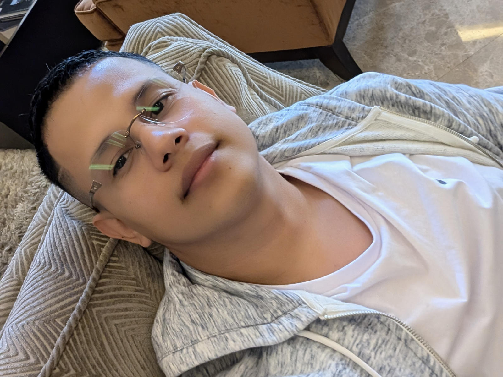 LiamB with glasses, short black hair, white t-shirt, gray hoodie, lying down, smiling slightly, wearing a black cap, on a textured beige surface.