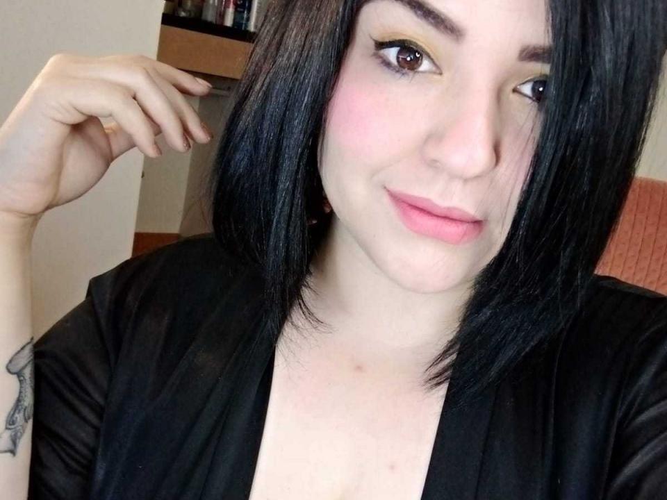 XimenaSexxyHot with black bob, light pink lipstick, subtle eyeliner, black top, tattoo on left forearm, slight blush, cute smile, hand near face, fair skin, black hair.