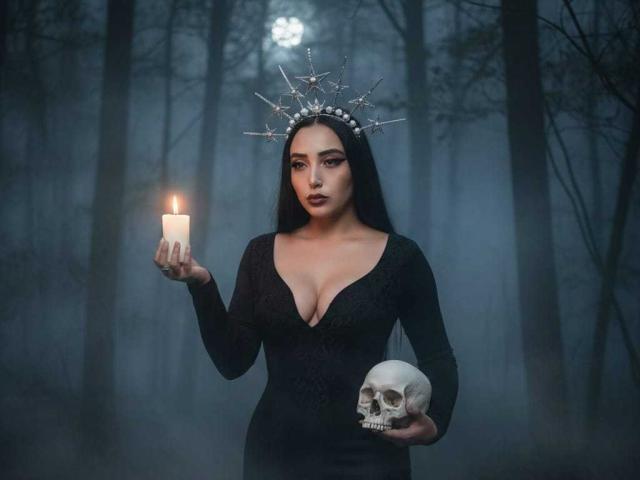 AmberGoddesss stands in a dark, misty forest with tall trees. She has long black hair, a deep cleavage in a black, lace-patterned dress, and wears a spiked headpiece. She holds a lit candle in her right hand and a skull in her left. The candle's flame is bright, contrasting with the dark, moody background. Her expression is serious, and her makeup is bold. The image has a gothic, mysterious vibe.