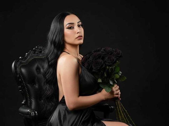 AmberGoddesss with long black hair, wearing a black satin dress, sitting on a black leather chair, holding a bouquet of dark purple roses, looking seductive.