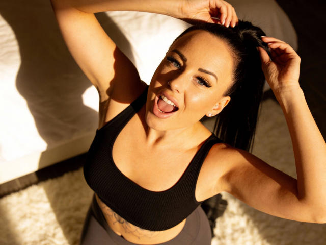 MilaJane-hot with black hair, smiling, tongue out, hands in hair, wearing black sports bra, exposing small tits, tattoo below, sitting on white rug, sunlight.