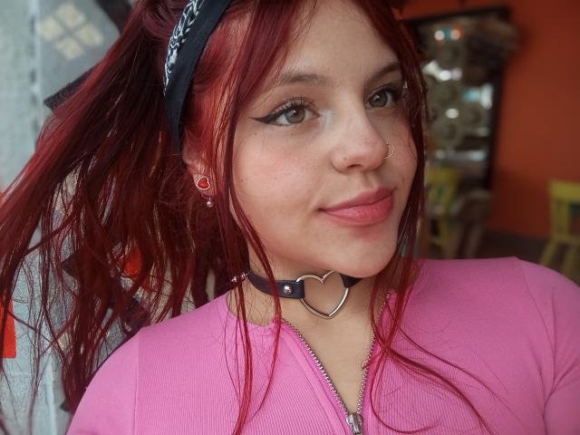 Sophie Hill, con i capelli rossi, una maglietta rosa, una collana nera con un cuore, un piercing al naso, eyeliner, orecchini rossi, un leggero sorriso, che guarda la fotocamera, sfondo sfocato, labbra rosa..