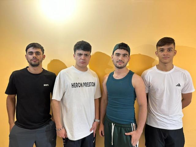 Four SpaceHotMen stand in a row, wearing black, white, teal, and white shirts with green shorts, looking serious, against a yellow wall. Their shirts are tight and muscular.