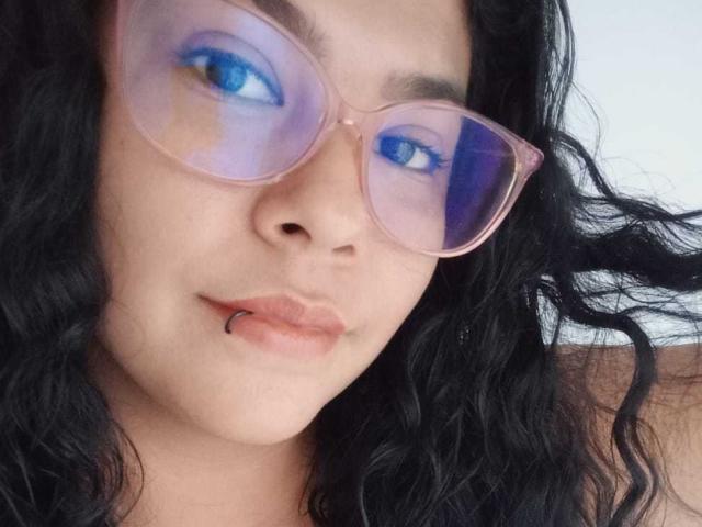DanielaBebe with blue eyeshadow, pink glasses, black wavy hair, and a black lip ring, looking at the camera. Her tits are not visible. She's got a cute face.