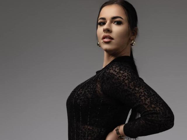 AndreaShow with dark hair, wearing a black lace dress, looking over her shoulder, showing her tits, wearing gold earrings, and a bracelet, standing against a gray background.