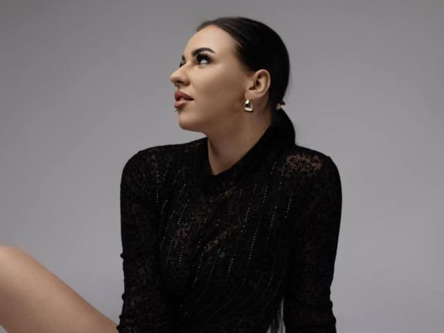 AndreaShow has dark hair in a ponytail, wearing a black lace top, looking to the side with a nose ring, and small earrings. Her tits are partially visible.