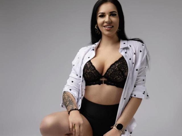 AndreaShow with black lace bra, showing her tits, wearing a white shirt open, black high-waisted shorts, and a tattoo on her right arm, smiling at the camera.