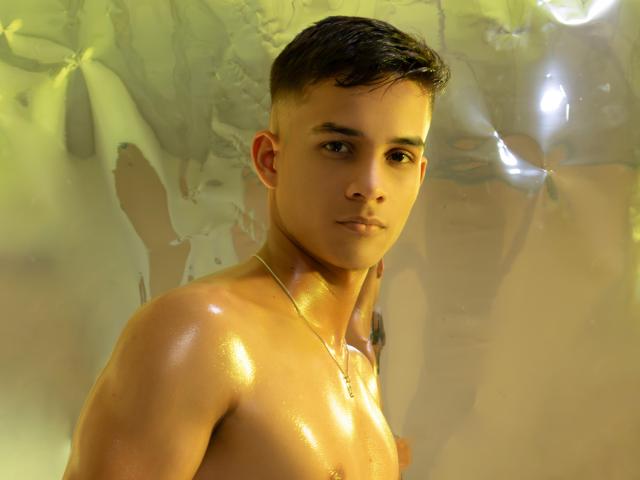 Shiny, oiled-up SpaceHotMen with short brown hair, looking at camera, has a smooth, muscular chest, and a gold necklace. Background is blurry with yellow-green light.