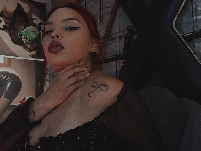 AlanaRoth has red hair, heavy black eyeliner, and red lipstick. She's wearing a black sheer top with small sparkles. Her left hand is touching her neck, showing a tattoo on her shoulder. Her right hand has a tattoo on her wrist. She has a lip ring and black nail polish. The background is dimly lit with a poster partially visible. The image has a dark, moody tone.