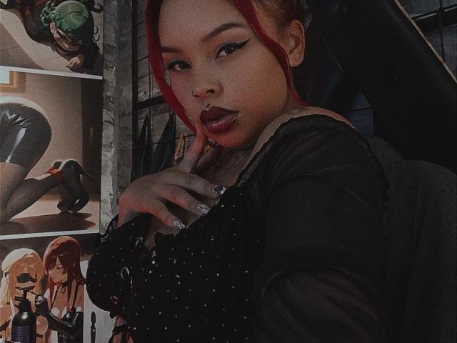 A photograph of AlanaRoth with red hair, wearing a black, sheer, long-sleeve top with sparkly details. She has dark lipstick, winged eyeliner, and a nose ring. Her left hand with glittery nails touches her chest. The background features anime-style posters with characters in black latex and red high heels. The image has a dark, moody tone.