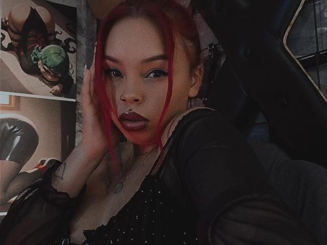 AlanaRoth with red hair, black sheer top, and nose ring, hand on face, looking at camera, showing tits and a small tattoo on her neck. Dark, moody selfie.