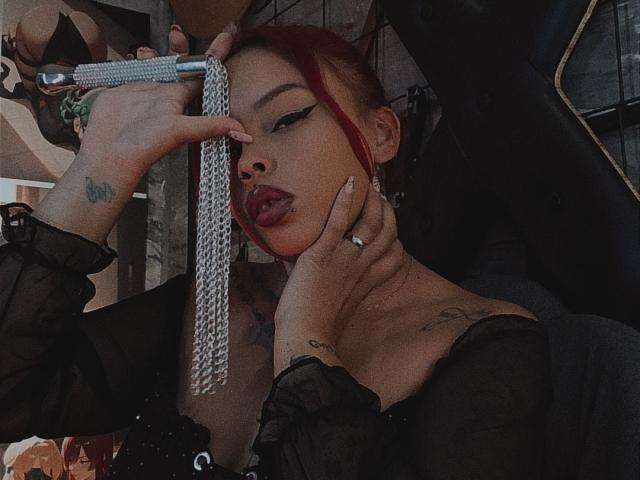 AlanaRoth with red hair, black off-shoulder top, holding a silver chain in her mouth, hand on her neck, tattoos visible, wearing ring, looking seductive, tongue out.