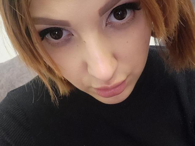 DinaHeart's face, short blonde hair, dark eyebrows, brown eyes with long eyelashes, wearing a black ribbed shirt, slight pout, soft skin, small beauty mark below right eye.