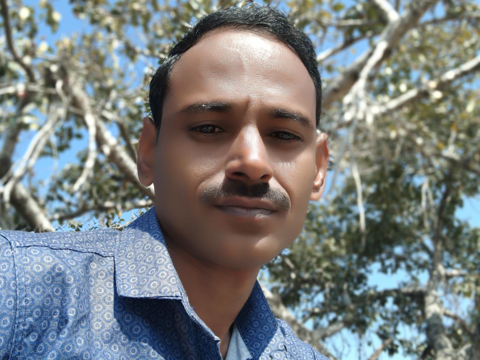 Indianbigdick with dark hair, mustache, and brown skin, wearing a blue patterned shirt, smiling slightly, standing outdoors with trees in the background, under bright sunlight.