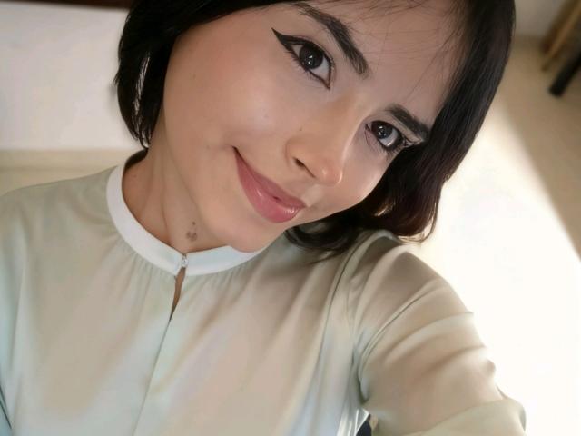 MariaFe has smooth light skin, black eyeliner, and short black hair. She's smiling with pink lips, wearing a light green shirt, and a small beauty mark near her chin.