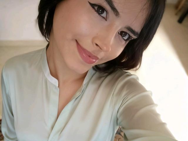 MariaFe with short black hair, light skin, and a cute smile, wearing a green button-up shirt, has small tits and is looking up at the camera.