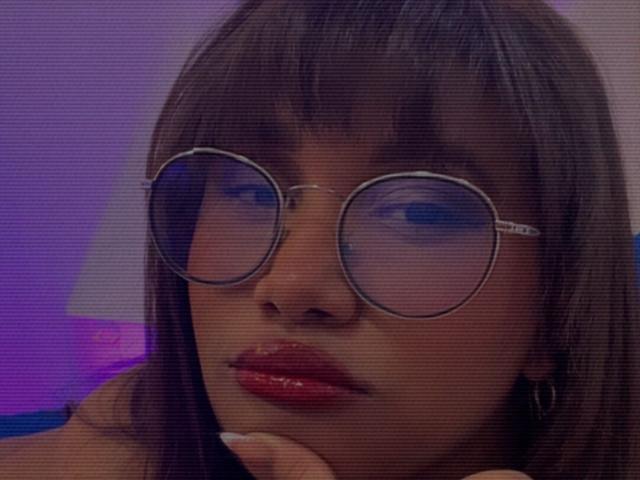 LoreDuval has dark brown, straight hair with bangs, wearing round glasses and red glossy lips, with a hand resting near her chin, in a purple-lit, slightly grainy photo.