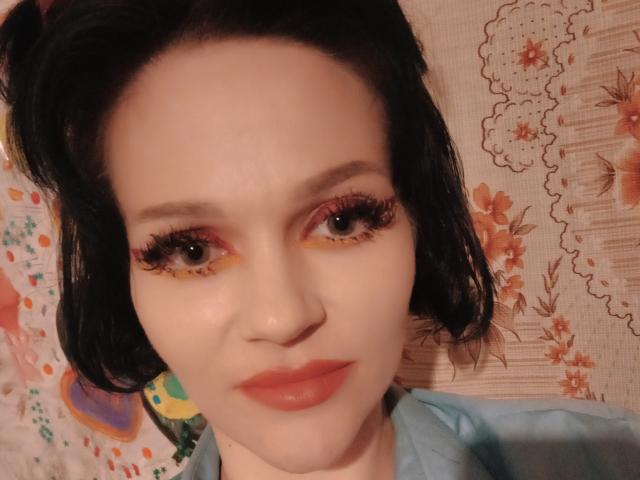 Farida69 with pale skin, black wavy hair, red eyeshadow, long eyelashes, orange lipstick, blue shirt, and a slight smile, looking directly at the camera.