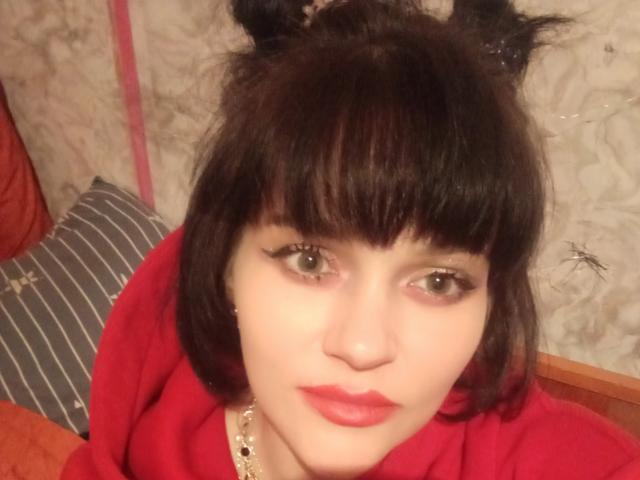 Close-up of Farida6, a young woman with pale skin, green eyes, and straight black hair with bangs. She wears a red top, bright pink lipstick, and a necklace with small beads. Her expression is neutral. She has black cat ears attached to her head. The background is blurry with a striped pillow on the left. The image has a casual, slightly rough quality.