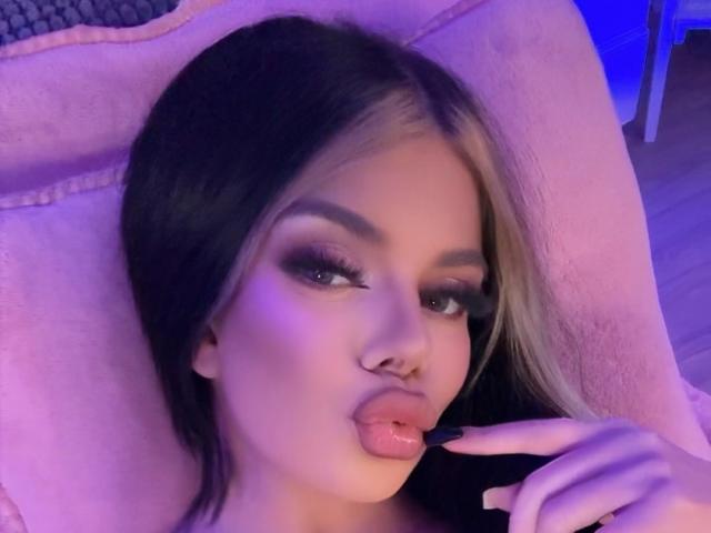 IvyRaven with pouty lips, blonde and black hair, dramatic eyeliner, and nose ring, sitting on pink chair, finger near mouth, blue and purple lighting, sultry expression.