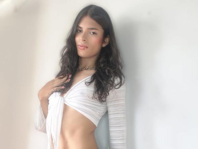 LunarisSmith with long black hair, wearing a white, ruched, long-sleeve top exposing her small tits, and a gold chain necklace, standing against a white wall.