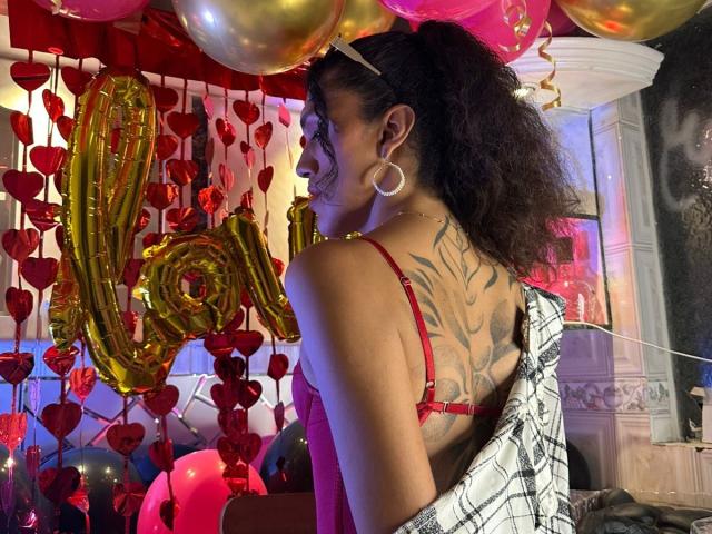 CrissOziz with a tattooed back, red bra, and curly hair, facing a wall of red hearts and gold balloons, wearing a plaid shirt pulled down.