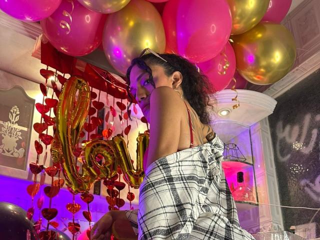 CrissOziz stands in a room with colorful balloons above, wearing a white and black plaid dress with red straps. Her curly brown hair is loose. She looks over her shoulder with a slight smile. The background has a metallic heart decoration and a large "LOVE" balloon. The room is illuminated with pink and purple lights. The composition focuses on CrissOziz and the vibrant balloon decor.