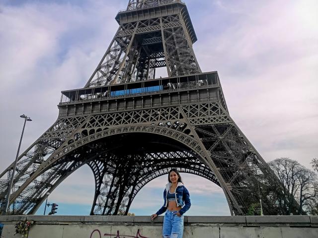 EmmaMae stands in front of the Eiffel Tower, wearing a blue denim jacket, white crop top, and high-waisted blue jeans. She's smiling with hands in pockets. The Eiffel Tower's intricate lattice structure dominates the background, with its arched supports visible. The sky is cloudy with soft pink and blue hues. EmmaMae's pose is relaxed, and her outfit is casual and stylish. The concrete barrier she stands on has graffiti.