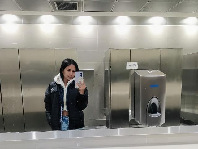 EmmaMae with black hair, wearing a black jacket and white hoodie, taking a mirror selfie in a metallic bathroom, showing her phone and a hand in her jacket.