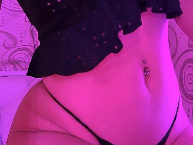 NaylaGrey's midsection with a black, polka-dotted top, black thong, and a jeweled navel piercing, under pink lighting. Her belly is soft, with visible skin folds.