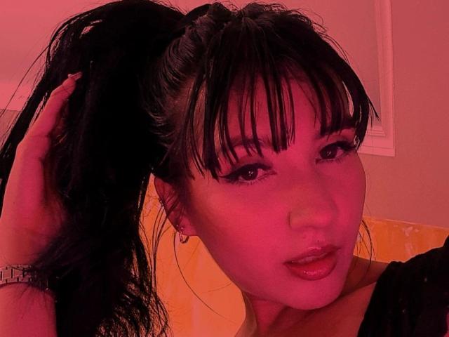 NaylaGrey tem cabelo preto em um rabo de cavalo alto, iluminação vermelho-rosa, franja e está de língua para fora. Sua mão está no cabelo, mostrando um relógio de prata..
