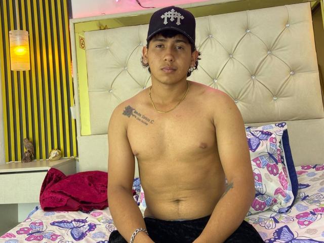AxelMonroe, shirtless, wearing a black cap with a white emblem, has a tattoo on his right shoulder, sitting on a bed with butterfly-patterned sheets, and a red pillow.